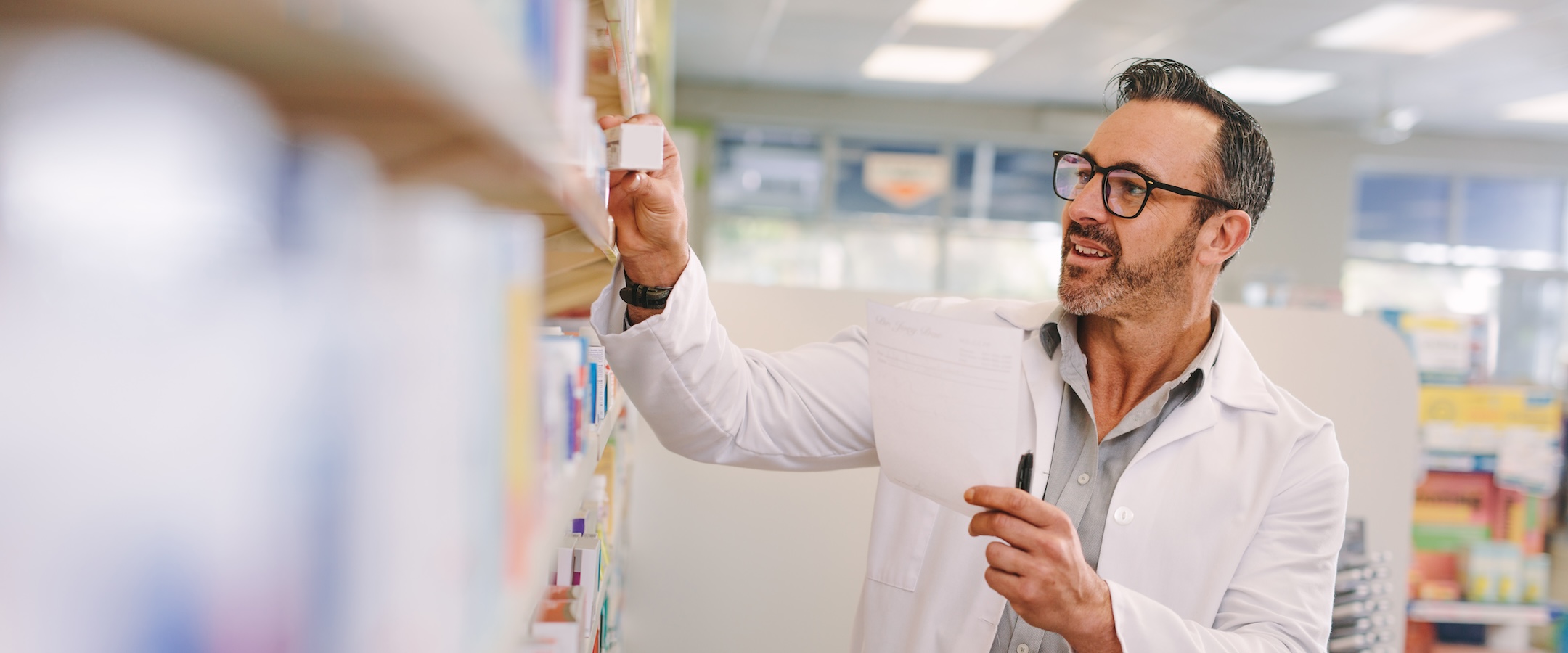 Mature chemist with a prescription searching right medicine on shelves in pharmacy. Male pharmacist holding prescription checking medicine in pharmacy.